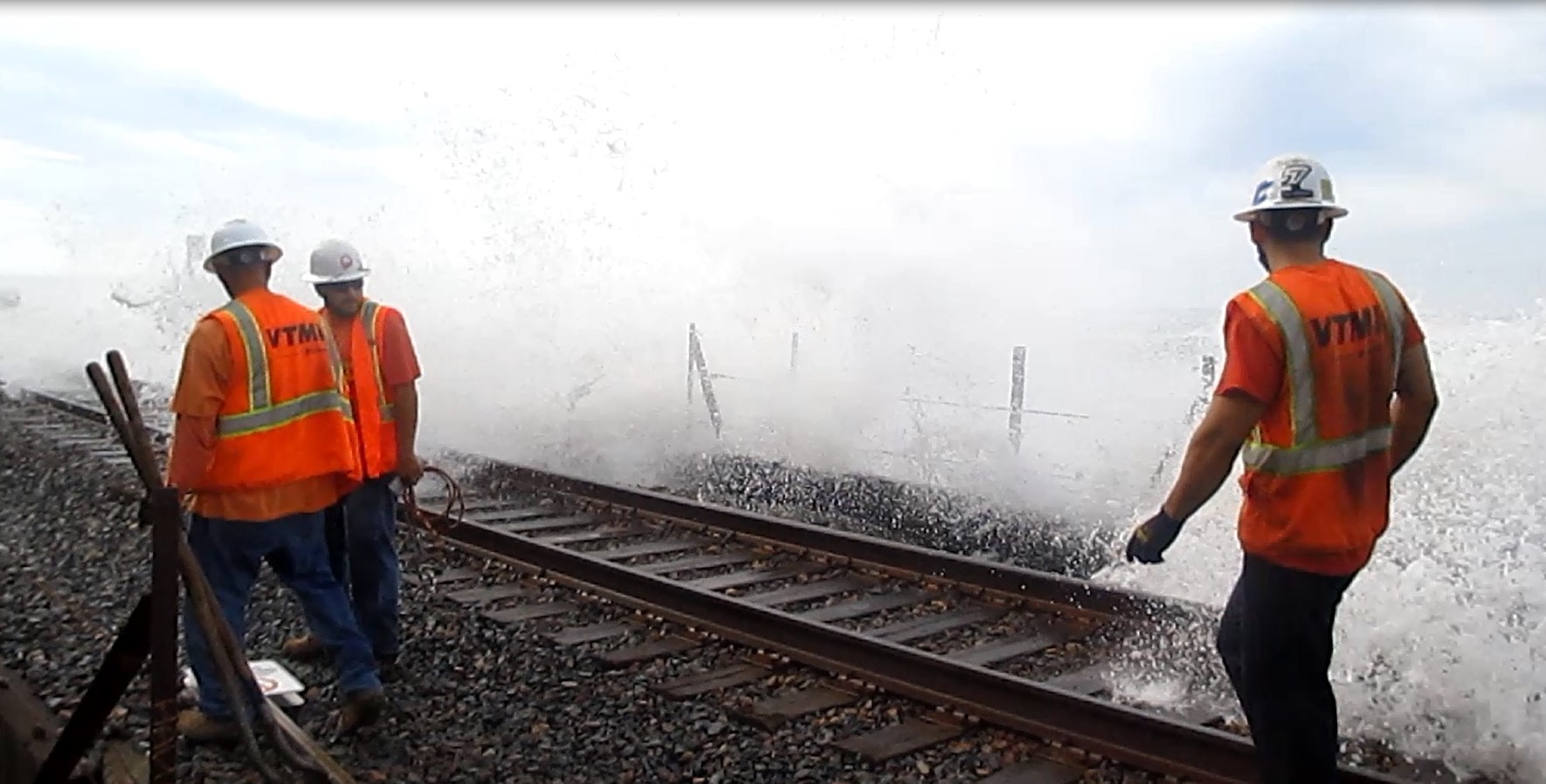 3 Caltrans workers standing on a rail line as waves overtop the rails.